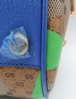 Close-up of a Gucci handbag corner showing a blue leather trim, brown and beige GG monogram canvas, and bright green fabric panels, with a gold-tone metal stud partially covered by blue tape.