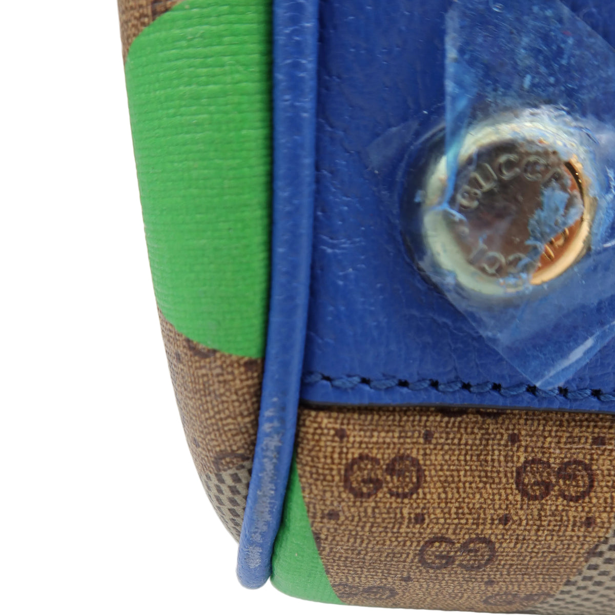 Close-up detail of a Gucci handbag corner showing the brown GG monogram canvas, a bright blue leather section, and green color-blocked panels, with a protective clear film covering part of the metal stud.