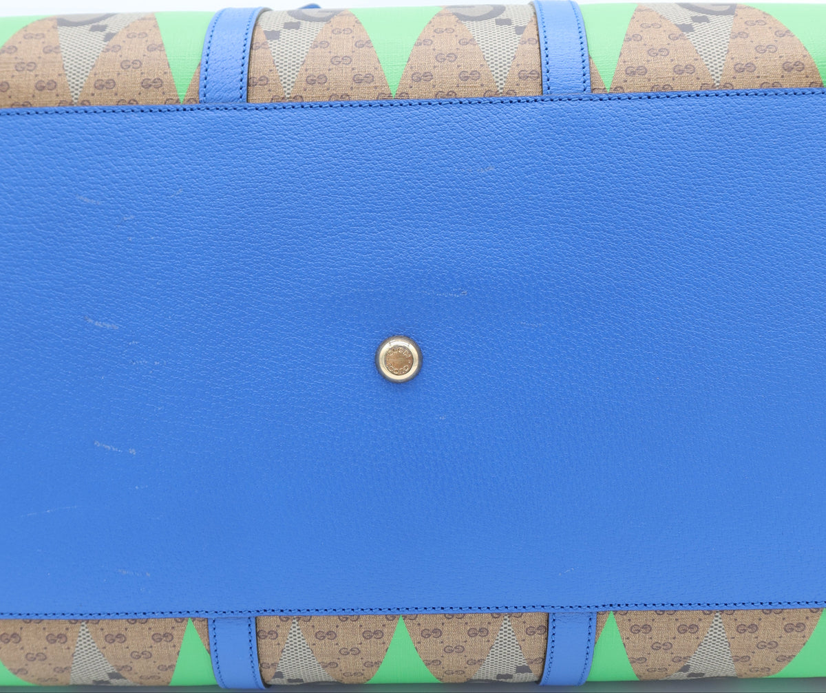 A close-up of a Gucci handbag showing the bottom flap, featuring a vibrant blue pebbled leather center with a gold-tone snap button, and green and beige GG Supreme patterned fabric borders on the top and bottom.