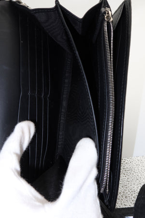 Close-up of the interior of a black leather clutch or wallet-style handbag, showing multiple card slots, a zip pocket, and a white gloved hand holding it open.