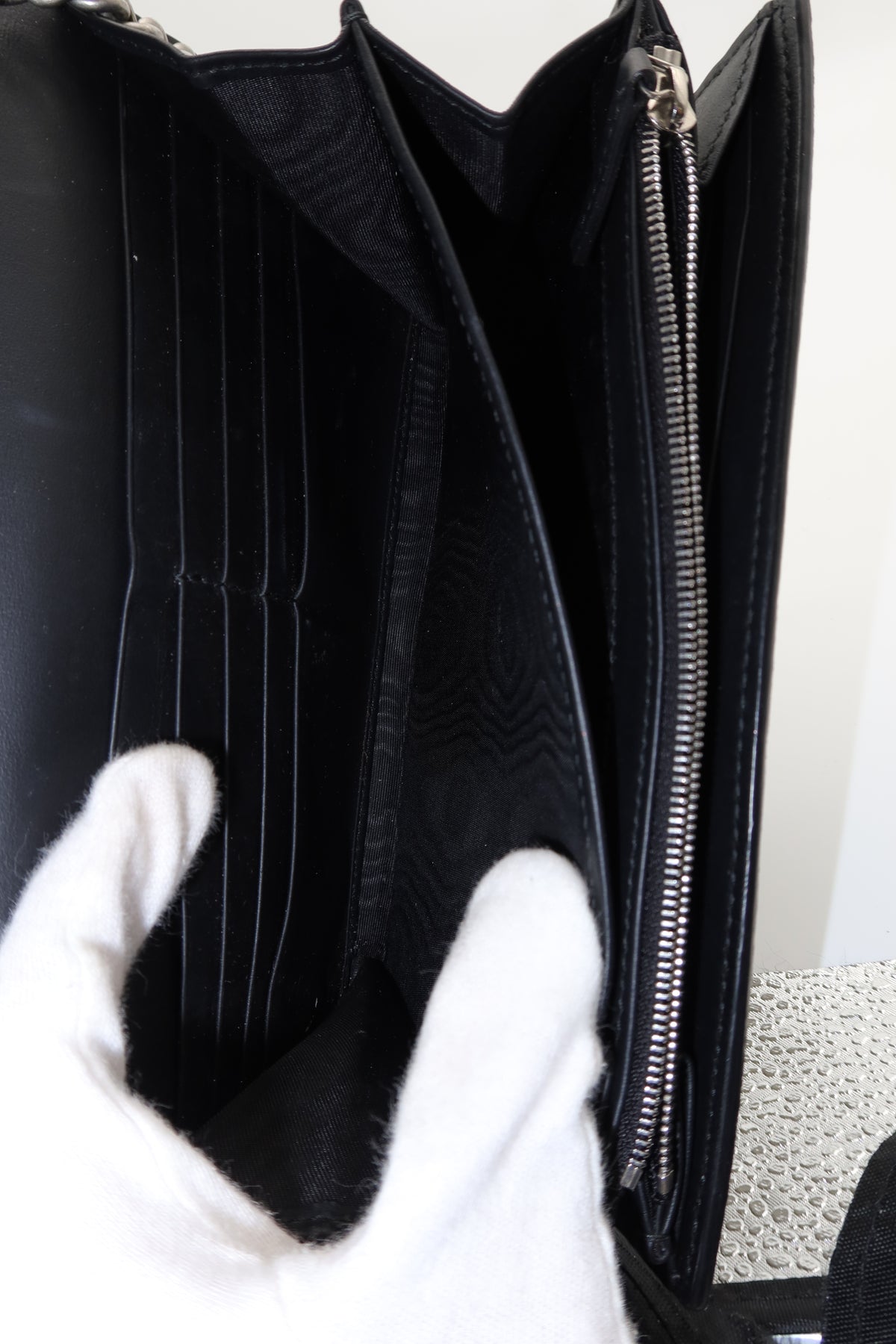 Close-up of the interior of a black leather clutch or wallet-style handbag, showing multiple card slots, a zip pocket, and a white gloved hand holding it open.