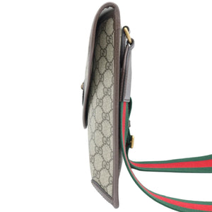 A Gucci monogram canvas crossbody bag shown from a side profile view, featuring the signature GG pattern in beige and brown, brown leather trim, and a green and red web stripe strap.