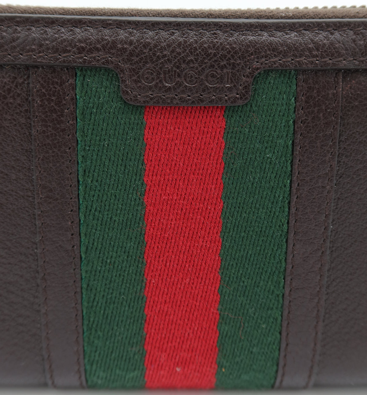 Close-up of a brown leather wallet featuring the signature green and red web stripe pattern on the front, with a subtle embossed Gucci logo on the leather tab above the stripes.